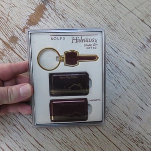 May include: A Rolfs Hideaway Spare Key Gift Set in a clear plastic box. The set includes a gold-tone key chain with a burgundy key holder, a burgundy spare key holder with a gold house design, and a burgundy magnetic spare key holder with a gold house design. The box has the words "Rolfs Hideaway Spare Key Gift Set" printed on it.