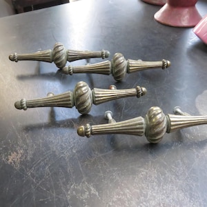 May include: Set of four antique brass drawer pulls. Each pull features a decorative, ribbed center section and tapered ends. The pulls are arranged on a dark surface, showcasing their aged patina and classic design.