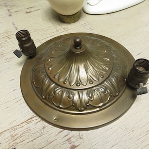 Puede incluir: Una lámpara de techo vintage de latón con dos portalámparas. La lámpara tiene un diseño floral en relieve ornamentado y un remate central. El metal tiene una pátina cálida y envejecida, lo que sugiere su antigüedad.