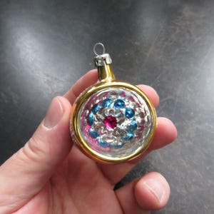 May include: A round, gold-colored Christmas ornament with a clear glass center. The center is decorated with a pink and blue design with a large pink jewel in the middle.