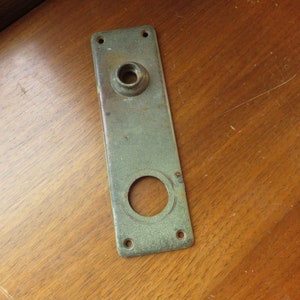 May include: A vintage, rectangular, brass door plate with a circular hole and a smaller circular hole with a raised lip. The plate has a greenish patina.