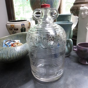 May include: A clear glass jug with a red cap and two handles. The jug has a decorative embossed pattern around the middle and horizontal ridges near the bottom. It is sitting on a dark surface.