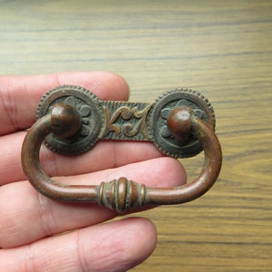 May include: Antique brass drawer pull with a decorative design and a ring handle. The pull has a dark patina and is in good condition.