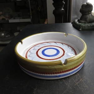 May include: A round ceramic ashtray with a white, blue, and red concentric circle design. The ashtray has a green and brown rim with a white and red stripe.