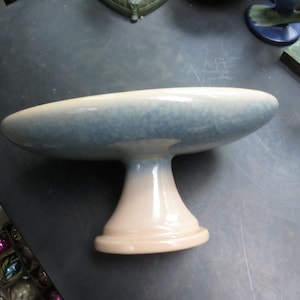 May include: A ceramic pedestal cake stand with a light blue and white glaze. The stand has a wide, shallow bowl and a tall, round base.