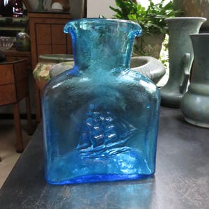May include: A blue glass pitcher with a textured surface and a ship design embossed on the front. The pitcher has a wide mouth and a narrow neck.