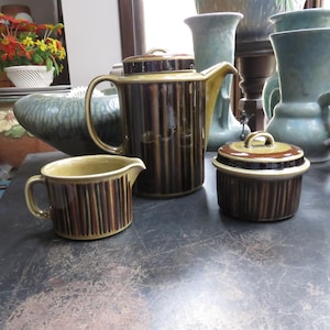 May include: A vintage brown and tan ceramic tea set with a creamer, sugar bowl, and teapot. The set has vertical striped detailing and a light tan rim. Other ceramic vases and bowls are in the background.