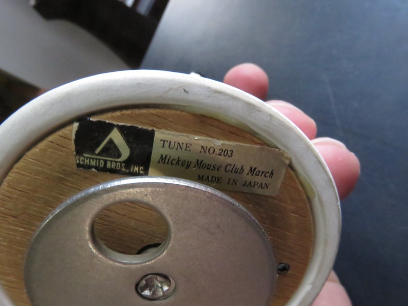 May include: Close-up of the bottom of a wooden music box with a white porcelain top. The music box plays "Mickey Mouse Club March" and is labeled "Tune No. 203" and "Made in Japan".