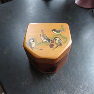 May include: Wooden hexagonal box with a light brown lid and a darker brown base. The lid features a painted design of four small birds perched on green branches with red berries. The box is on a dark surface.