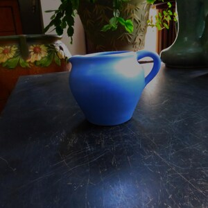 May include: A light blue ceramic creamer or pitcher with a rounded body and a handle. The creamer is sitting on a dark wooden surface.