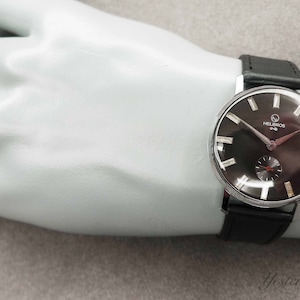 May include: A silver wristwatch with a black face and black leather band. The watch face has white markings and the text "Helbros One" printed on it.