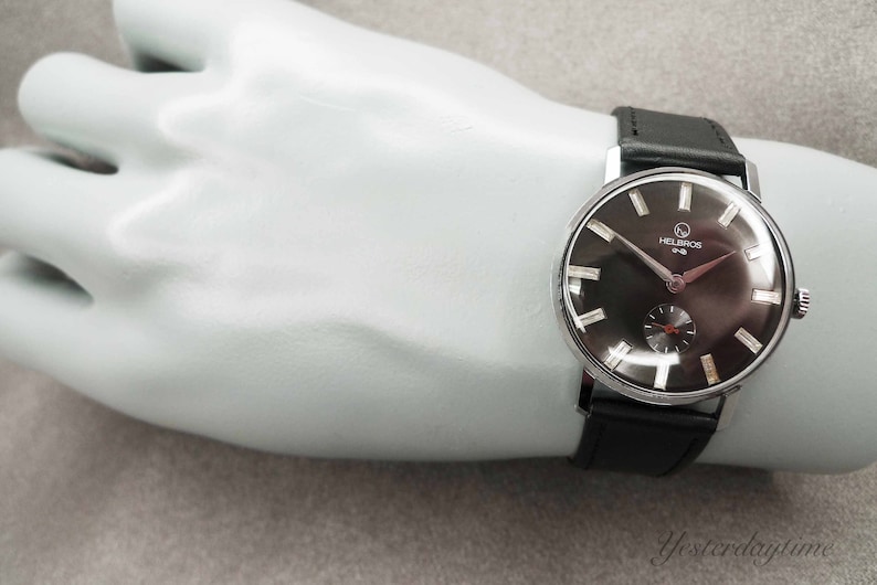 May include: A silver wristwatch with a black face and black leather band. The watch face has white markings and the text "Helbros One" printed on it.