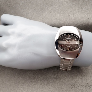 Puede incluir: Un reloj de pulsera automático Zenith Defy plateado con esfera gris oscuro y visualización de la fecha. El reloj tiene una caja rectangular y una correa de eslabones plateada. La palabra "Zenith" es visible en la esfera.