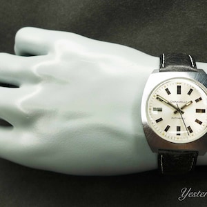 Pode incluir: Um relógio de pulso automático Caravelle prateado com mostrador branco e pulseira de couro preta. O relógio tem uma caixa quadrada e é usado no pulso de um manequim.