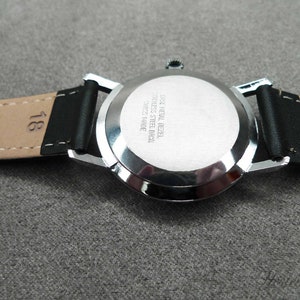 May include: Back view of a silver wristwatch with a black leather band. The watch face has the text "Case Metal Bezel Stainless Steel Back Swiss Made" engraved on it.