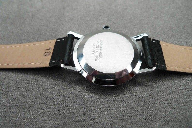 May include: Back view of a silver wristwatch with a black leather band. The watch face has the text "Case Metal Bezel Stainless Steel Back Swiss Made" engraved on it.