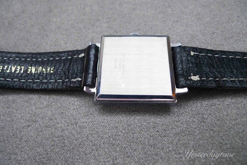 May include: A silver square watch with a black leather strap. The strap has white stitching and the text "Genuine Leather" is printed on the strap.