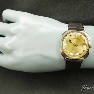 May include: A gold-toned wristwatch with a square face and a brown leather band. The watch face has black Roman numeral hour markers and a date window at the 3 o'clock position. The watch face also has the text "Automatic" and "21 Jewels" printed on it.