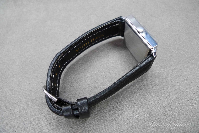 May include: A silver square-shaped wristwatch with a black leather band. The band is labeled "Genuine Leather" on the back.