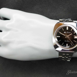 Puede incluir: Un reloj de pulsera plateado con una esfera negra y agujas plateadas. La esfera del reloj tiene el texto "K-time" y "21 Jewels" impreso en ella.