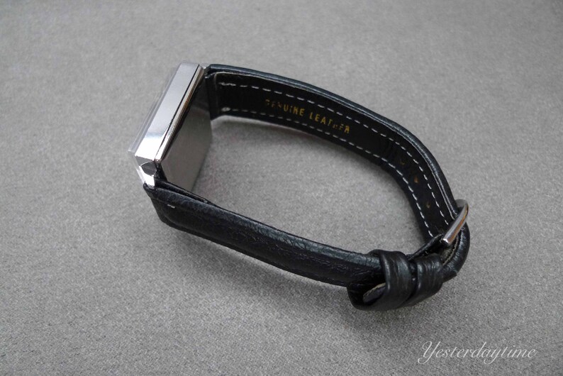 May include: A silver square watch with a black leather band. The band is stitched with white thread and has the words "Genuine Leather" printed on it.