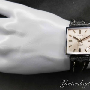 May include: A silver square-shaped wristwatch with a white face and black leather band. The watch face has black hands, black Roman numeral hour markers, and the text "GROK" and "17 JEWELS INCABLOC".