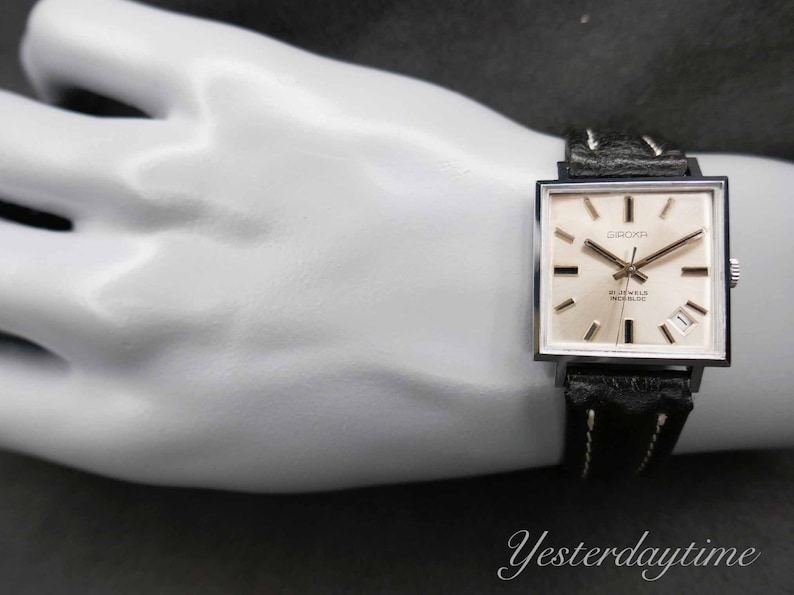 May include: A silver square-shaped wristwatch with a white face and black leather band. The watch face has black hands, black Roman numeral hour markers, and the text "GROK" and "17 JEWELS INCABLOC".