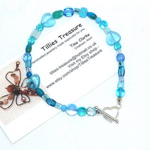 May include: A blue and green beaded bracelet with a heart-shaped clasp. The bracelet is handmade and features a variety of blue and green beads, including some heart-shaped beads. The bracelet is on a white background.