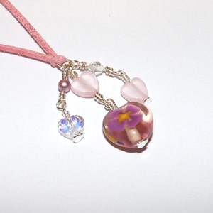 May include: A pink suede cord necklace with a pink glass heart pendant featuring a purple flower. The pendant is accented with smaller pink heart charms and clear crystal beads.