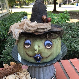 May include: A green pumpkin with a painted face, wearing a witch's hat with a brown brim and a tattered white fabric. The pumpkin is sitting on a silver pedestal with a decorative edge. Two small red and black skull-shaped ornaments are on the hat.