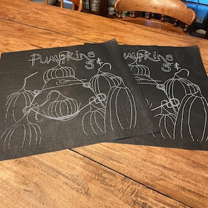 May include: Two square black fabric placemats featuring a white line-art illustration of multiple pumpkins. The text "Pumpkins 5¢" is printed in a stylized white font at the top of each mat. The design uses simple white outlines on a dark background.