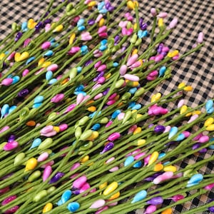 Op de afbeelding: Een bundel kunstmatige bloemstelen met groene stelen en kleurrijke, druppelvormige kralen in geel, blauw, roze en paars. De stelen zijn diagonaal gerangschikt tegen een geruite achtergrond, wat een levendige decoratie creëert.