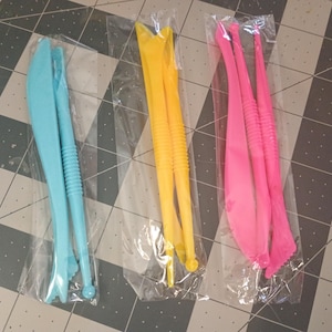 May include: Three sets of plastic clay sculpting tools in clear plastic packaging. The tools are in three colors: light blue, yellow, and pink. Each set contains two or three tools with different shaped ends and textured handles.