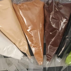 Könnte beinhalten: Sieben Plastiktüten, gefüllt mit farbigen Pulvern, fächerförmig angeordnet. Die Farben sind Weiß, Beige, Braun, Schwarz und Grün. Die Tüten sind versiegelt und wahrscheinlich für Bastelarbeiten oder zum Backen bestimmt.