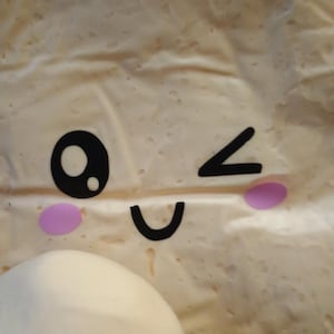 May include: A close-up of a light-colored, textured surface with a cartoon face design. The face includes a black eye, a winking eye, a curved mouth, and two pink cheek circles. A white, rounded object is in the lower left corner.