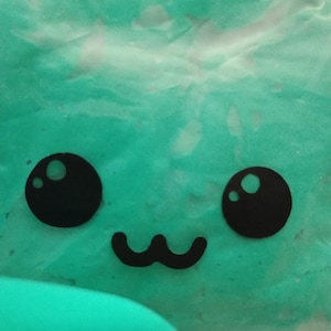 May include: Close-up of a teal-colored, textured surface with a cartoon face. The face features two large black circles for eyes and a curved black line for a mouth. The overall design is simple and playful.