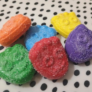 Może przedstawiać: Asortyment kolorowych, posypanych cukrem cukierków w kolorze pomarańczowym, niebieskim, czerwonym, zielonym, żółtym i fioletowym. Cukierki mają teksturowany wygląd i są ułożone na białej powierzchni z czarnymi kropkami. Słodka przekąska.