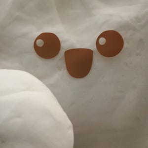 May include: A close-up of a white, fluffy object with a cartoon face. The face features two brown circular eyes with white highlights and a brown, curved mouth. The background is a soft, white texture.