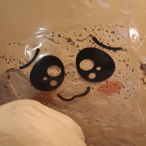 May include: A close-up of a tan-colored surface with a cartoon face sticker applied. The sticker features large black eyes, curved eyebrows, and a smiling mouth. The cheeks are a light pink color.