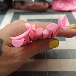 May include: A collection of pink plastic hair styling tools, held in a hand with a butterfly tattoo. The tools have various shapes, including rounded and petal-like designs. The person's fingernails are painted yellow.