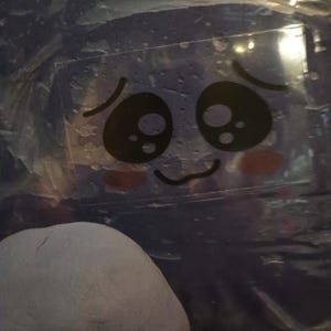 Könnte beinhalten: Ein klarer, rechteckiger Aufkleber mit einem Cartoon-Gesicht, das große schwarze Augen, rosige Wangen und einen gebogenen Mund aufweist. Der Aufkleber befindet sich auf einer blauen Oberfläche, mit einem abgerundeten, grauen Objekt im Vordergrund.