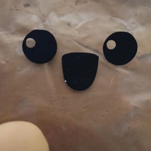 May include: A close-up of a face made from black cutouts. Two circles with white dots form the eyes, and a curved shape forms the mouth. A light-coloured, round object is in the lower left corner.