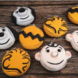 May include: A collection of ceramic ornaments featuring cartoon character faces. The ornaments are round and painted in various colors, including white, yellow, and black. Each ornament has a silver string for hanging, perfect for holiday decor.