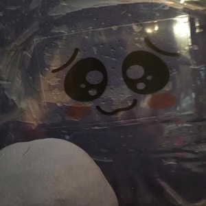 Könnte beinhalten: Ein klarer, rechteckiger Aufkleber mit einem Cartoon-Gesicht mit großen schwarzen Augen, einem kleinen gebogenen Mund und rosigen Wangen. Der Aufkleber befindet sich auf einer blauen Oberfläche, mit einem abgerundeten, grauen Objekt im Vordergrund.