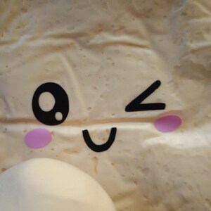 May include: A close-up of a light-colored, textured surface with a cartoon face design. The face includes a black eye, a winking eye, a curved mouth, and two pink cheek circles. A white, rounded object is in the lower left corner.