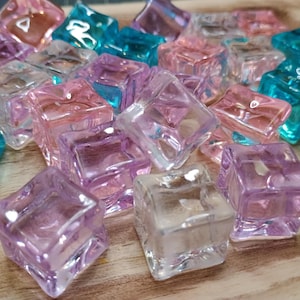 Puede incluir: Colección de gemas decorativas cuadradas, transparentes y coloridas en tonos rosa, morado y turquesa. Las gemas están dispersas sobre una superficie de madera marrón claro.