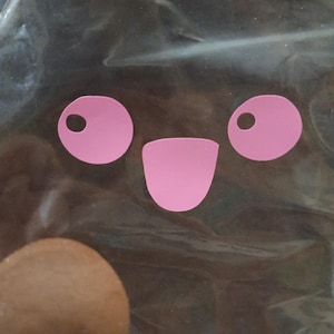 May include: Pink die-cut shapes arranged to resemble a smiling face. Two circles with smaller circles inside form the eyes, and a curved shape forms the mouth. The shapes are on a dark background.
