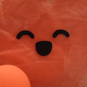 May include: Orange background with a smiling face made of black shapes. The face has two curved lines for eyes and a curved shape for a mouth. A round orange object is in the lower left corner.