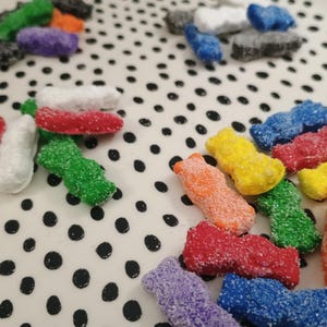 Może przedstawiać: Kolekcja kolorowych, cukrowych cukierków w różnych kształtach i kolorach, w tym czerwonym, zielonym, żółtym, pomarańczowym, fioletowym, niebieskim i białym. Cukierki ułożone są na białej powierzchni z czarnymi kropkami.