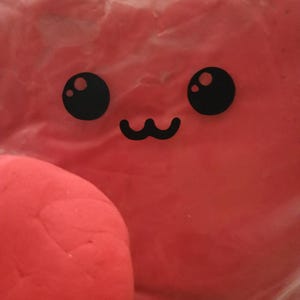 May include: A close-up of a red, heart-shaped object with a kawaii-style face. The face features two large, black, round eyes and a small, black, curved mouth. The object is in a clear plastic bag.